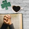 St Patrick's Breastplate Shamrock Ornament on wood surface next to heart-shaped coffee mug and Bible with hands clasped on top
