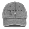 A gray, distressed baseball cap featuring the phrase "king of my heart" embroidered in black text across the front. Below the text are three simple symbols: a black crown, a white heart, and a black Christian fish symbol