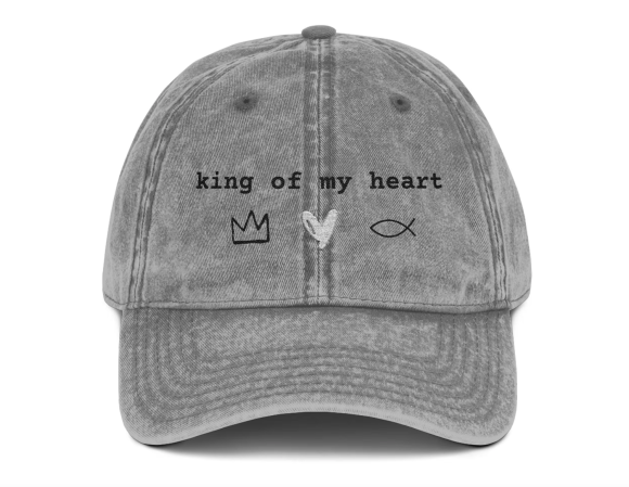 A gray, distressed baseball cap featuring the phrase "king of my heart" embroidered in black text across the front. Below the text are three simple symbols: a black crown, a white heart, and a black Christian fish symbol